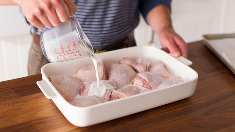 Raw chicken parts in a flat dish with high sides having buttermilk poured over them from a measuring cup