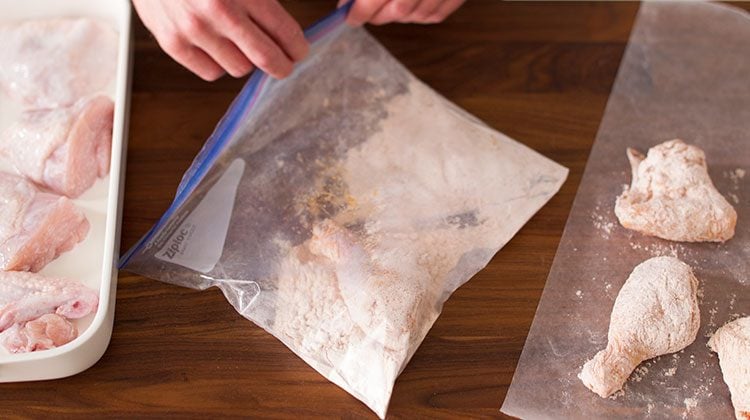 A person taking a drumstick from its buttermilk bath and getting it covered in coating inside a large resealable plastic bag then placing it on parchment paper to their left