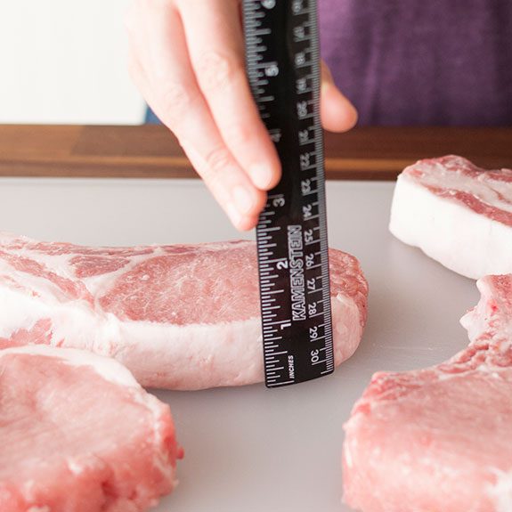 Person holding up a ruler to a raw pork chop to measure its thickness