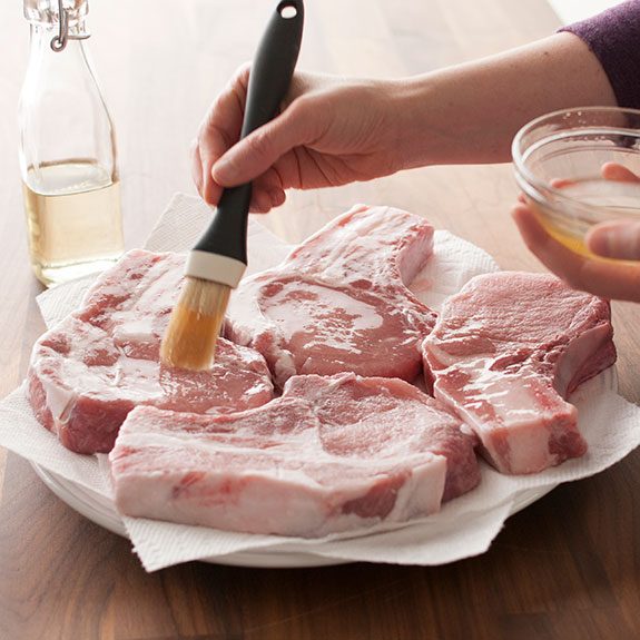 Person brushing oil over raw pork chops nestled together on a paper towel-lined plate