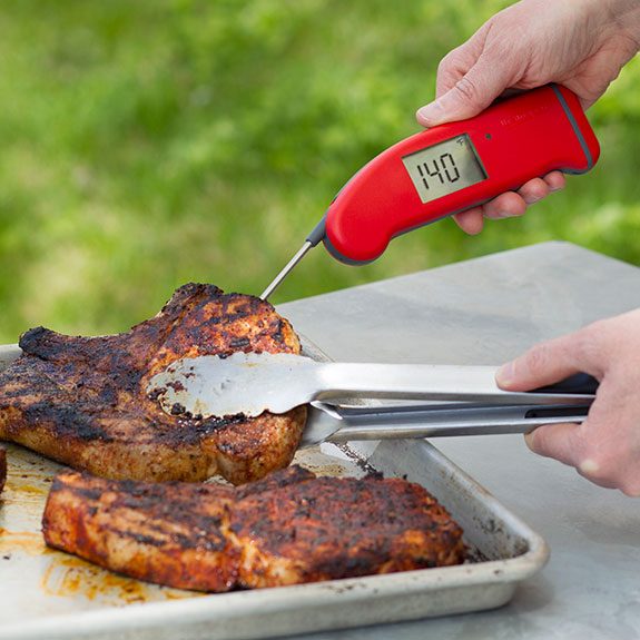 Person holding up a grilled pork chop from a baking sheet with metal tongs for a better angle for their red thermometer that is reading 140 degrees