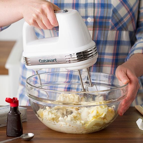 Person just beating the ingredients in a glass bowl with a hand mixer with a small container of vanilla extract nearby