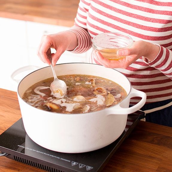 Person using a spoon to skim the foam off their broth from their dutch oven to put into a small glass cup