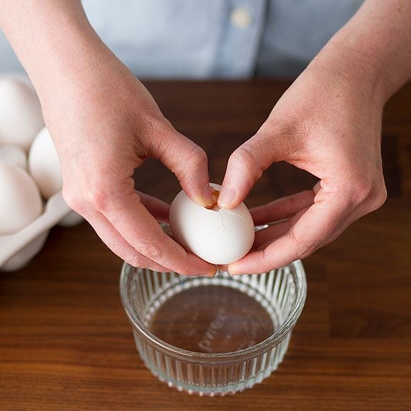Person digging their thumbs into the center of an egg that they're holding over a small glass cup
