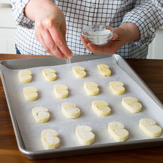 person sprinkling sugar over uncooked palmiers on a baking sheet