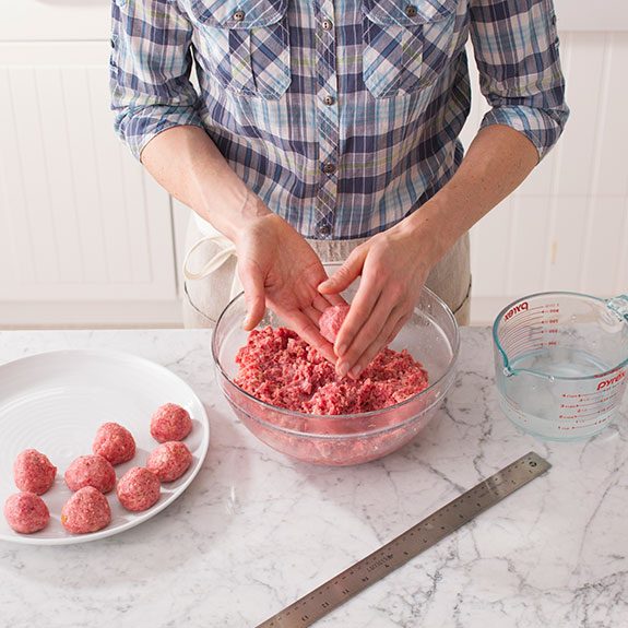 Person using their hands to roll the meat mixture from the glass bowl into balls that they then place on a separate white plate