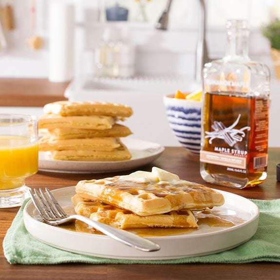 Two waffles covered in syrup and topped with butter sitting in front of a stack of waffles, glass of orange juice and a mostly full bottle of maple syrup