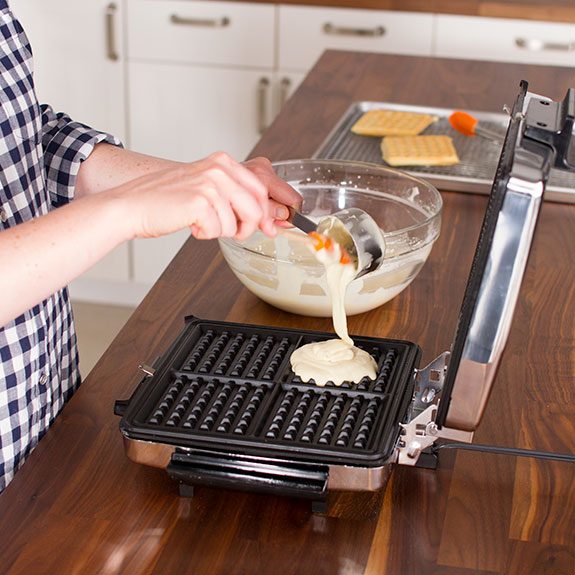 The mixture is being carefully poured into a waffle iron using a metal measuring cup