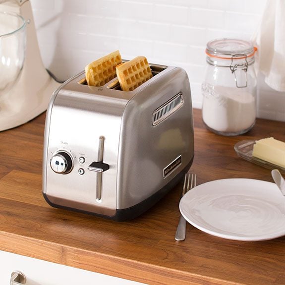 Two waffles sitting in the slots of a toaster with a plate and silverware ready beside the appilance